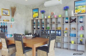 A dining area with a wooden table and wicker chairs, a whiteboard, and a shelf with various items. - White Knot Khao Yai (Pak Chong)