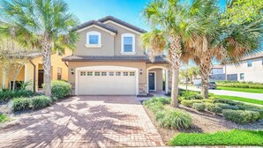 A two-story house with a garage, surrounded by palm trees and well-maintained landscaping. - Amazing 6Bd W/ GameRoom & Pool (Kissimmee)
