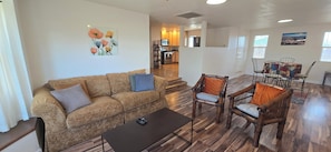 A living room with a sofa, coffee table, and chairs, a kitchen in the background, and artwork on the walls.