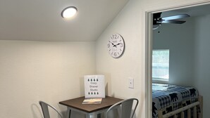 Ett litet rum med ett bord, två stolar, en klocka och en säng med ett rutigt täcke.