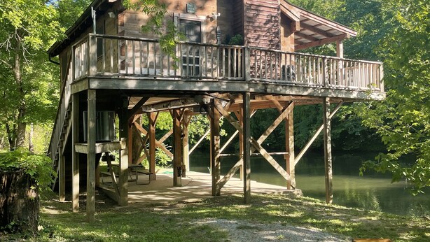 A wooden cabin with a deck, situated by a lake, surrounded by trees.