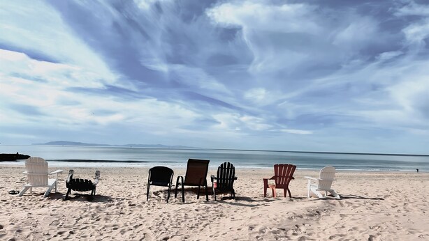 Une plage avec plusieurs chaises Adirondack et une table, donnant sur une mer calme sous un ciel nuageux.