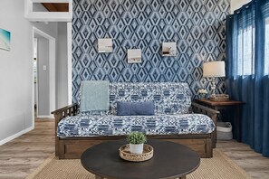 A cozy living room with a patterned sofa, a small table with a plant, and a wall with geometric wallpaper. - Cozy Beach View Loft, 42 Steps to the Sand + AC (Ventura)