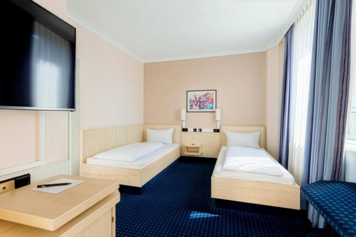 A hotel room with two single beds, a desk, a TV, a small bedside table, and a painting on the wall.