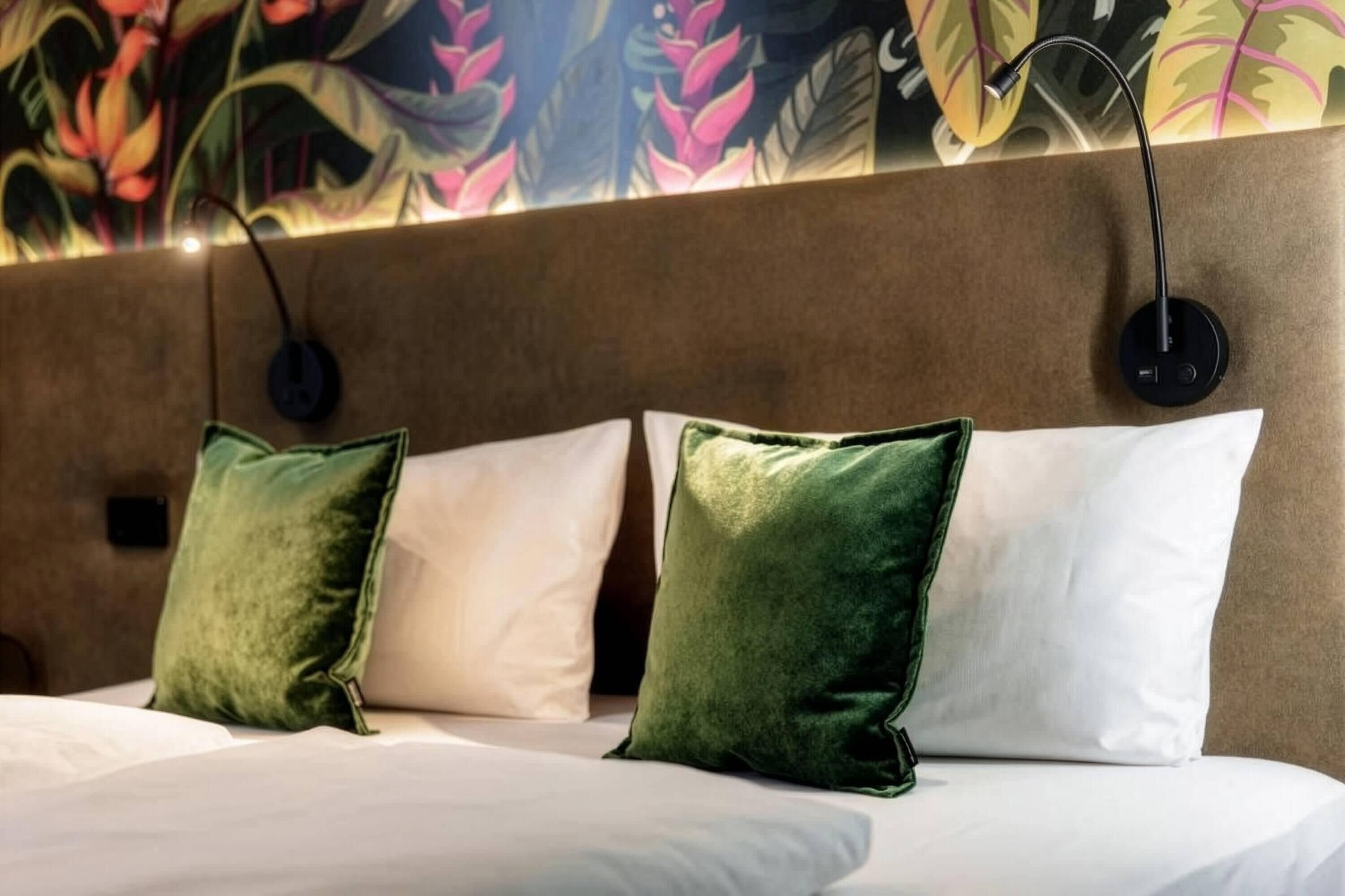Ein Bett mit weißen und grünen Kissen, ein Kopfteil mit floralem Wandbild und eine runde Wandleuchte.