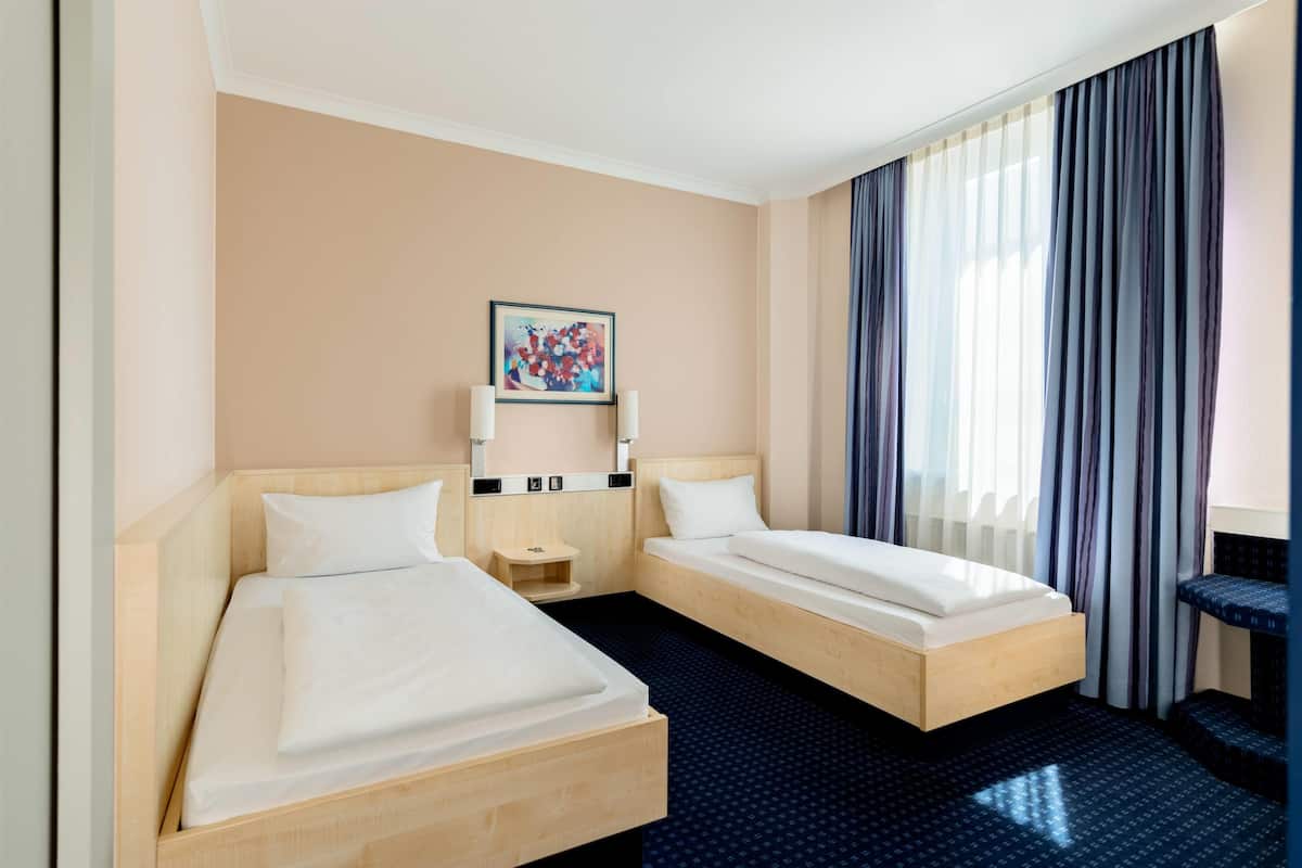 A hotel room with two single beds, a small bedside table, a nightstand, a wall-mounted TV, and a large window with curtains.