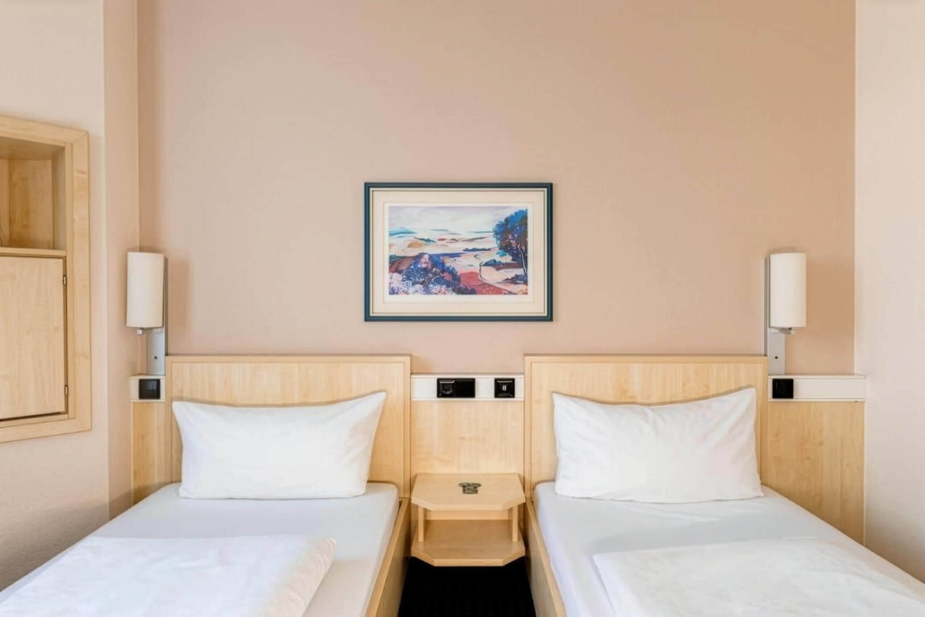 Een hotelkamer met twee eenpersoonsbedden, een klein nachtkastje met een lamp, een aan de muur hangend schilderij en een houten garderobe.