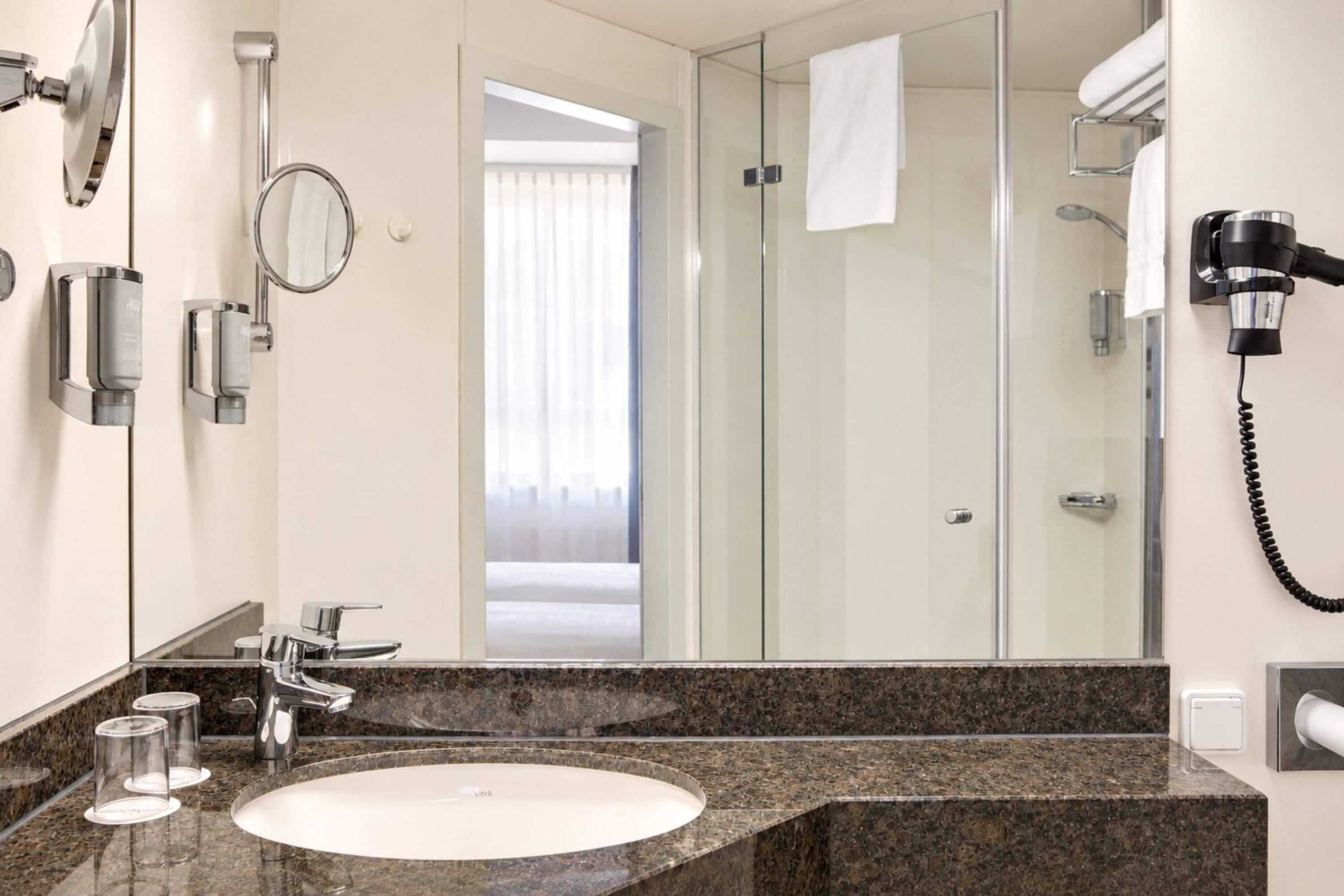 A modern bathroom with a marble sink, a handheld showerhead, a hairdryer, and a glass shower enclosure.
