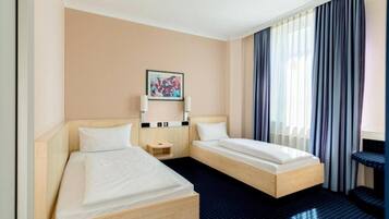 A hotel room with two single beds, a small bedside table, a nightstand, a framed picture on the wall, and a window with curtains.