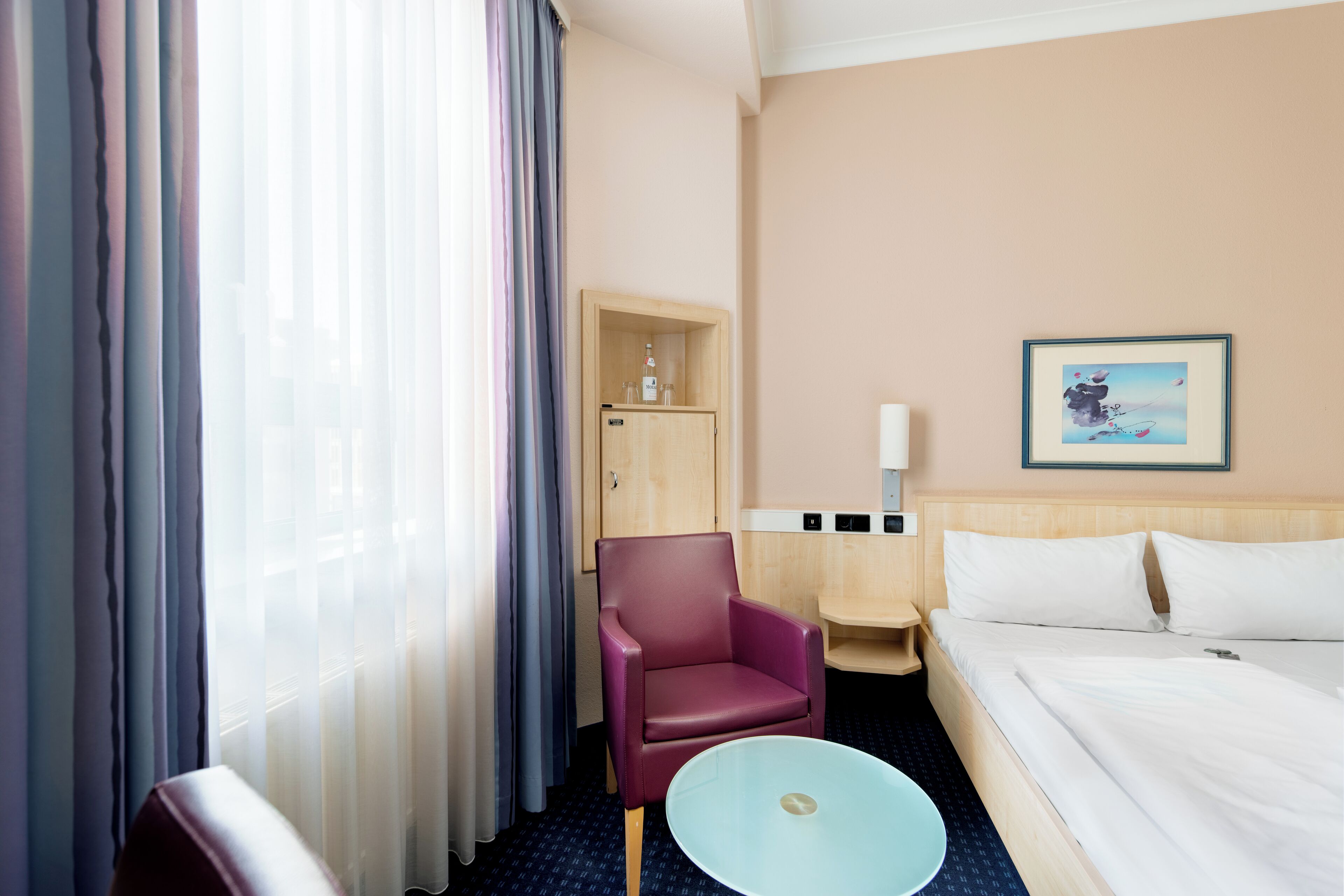 Habitación de hotel con cama, una mesita, una silla, una mesita de noche y un cuadro colgado en la pared.