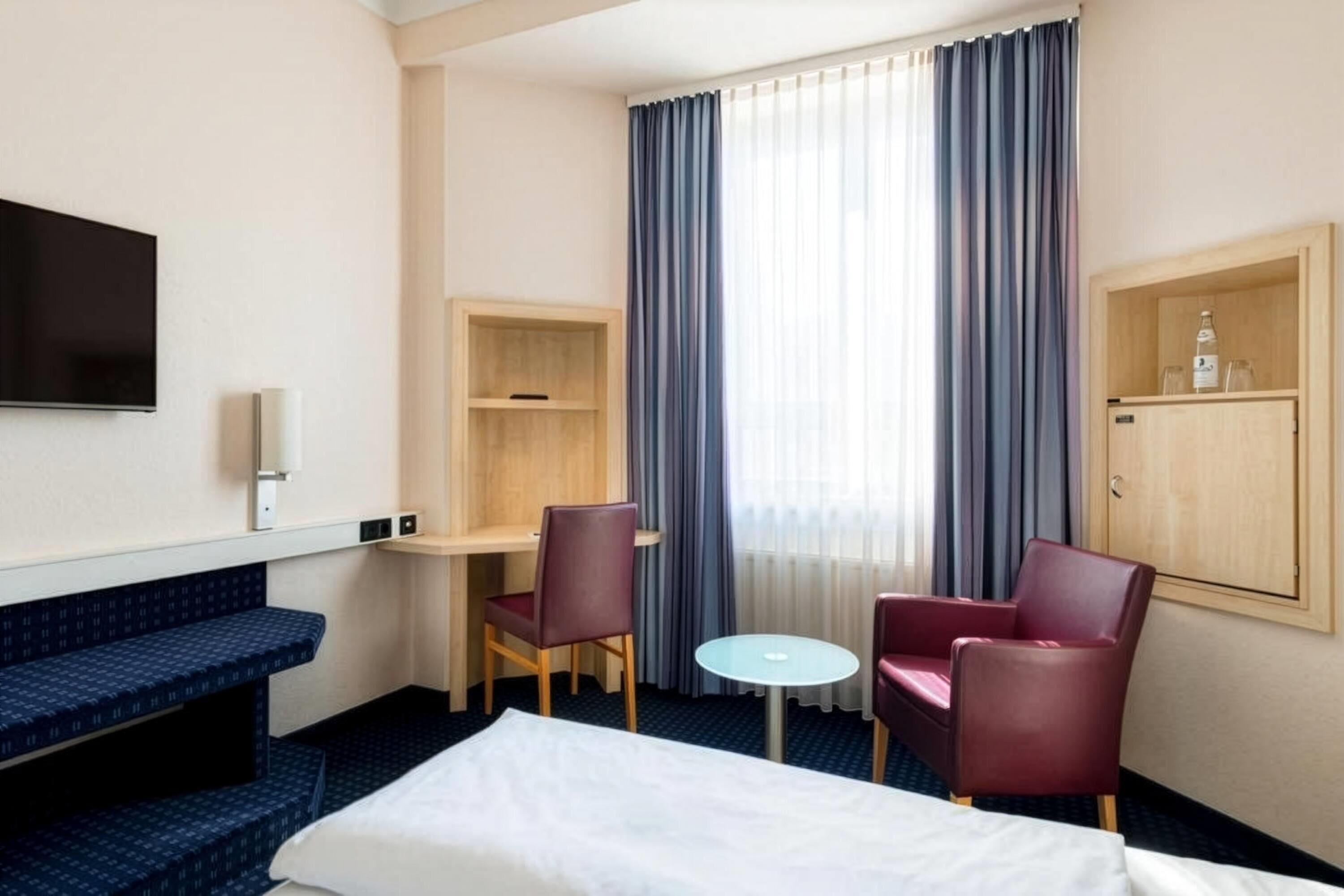A hotel room with a bed, a desk, two chairs, a TV, and a minibar.
