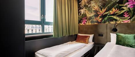 Habitación de hotel con cama, ventana con cortinas, cabecera con un mural floral y vistas a edificios.
