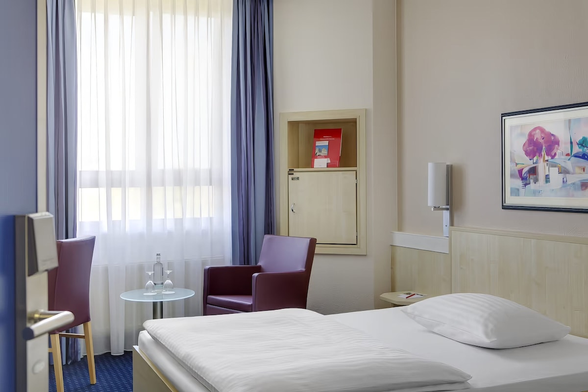 Una habitación de hotel con una cama, una silla, una mesita, un armario y un cuadro en la pared.