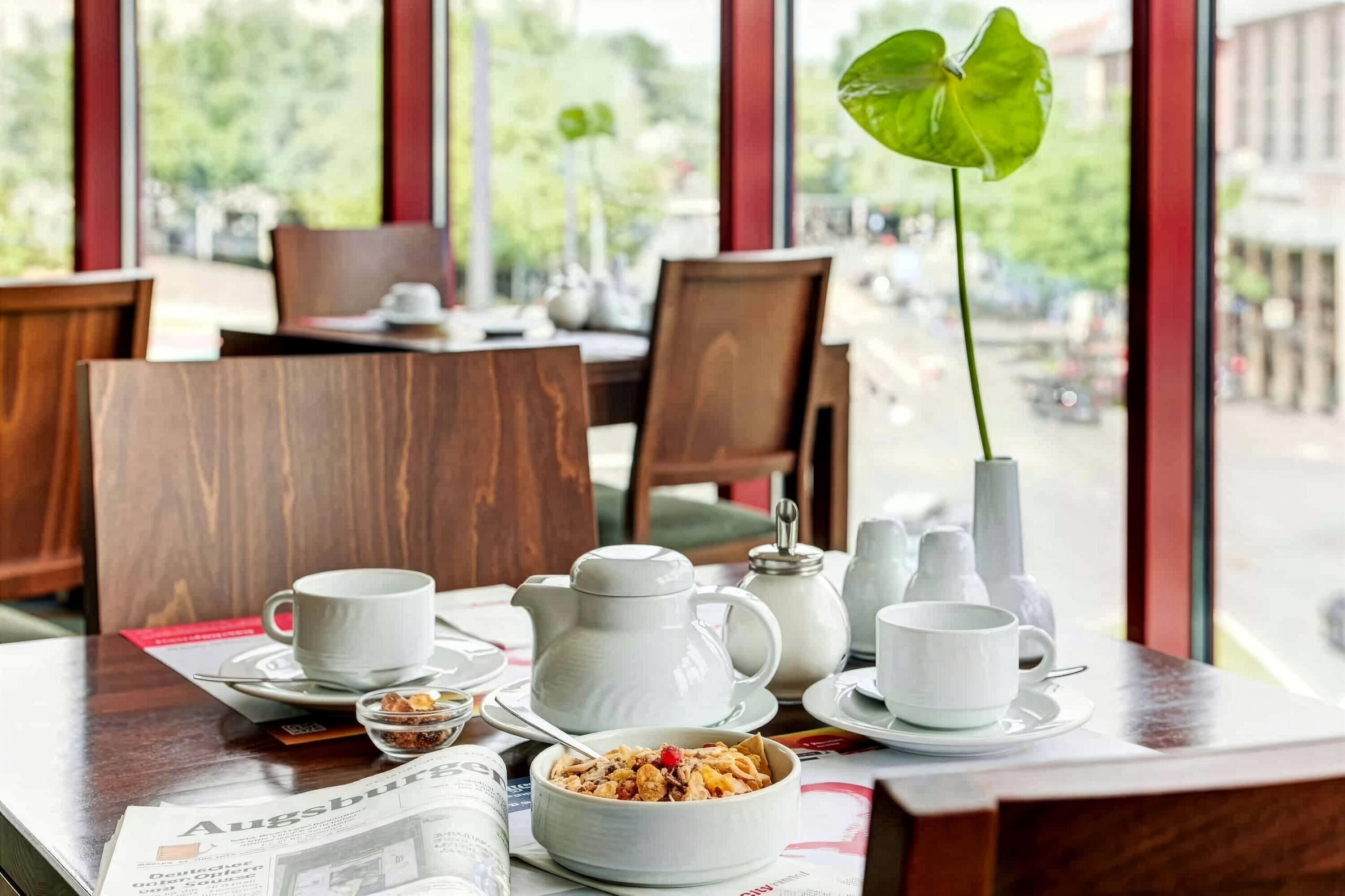 A dining table with white ceramic tableware, a bowl of cereal, and a newspaper.
