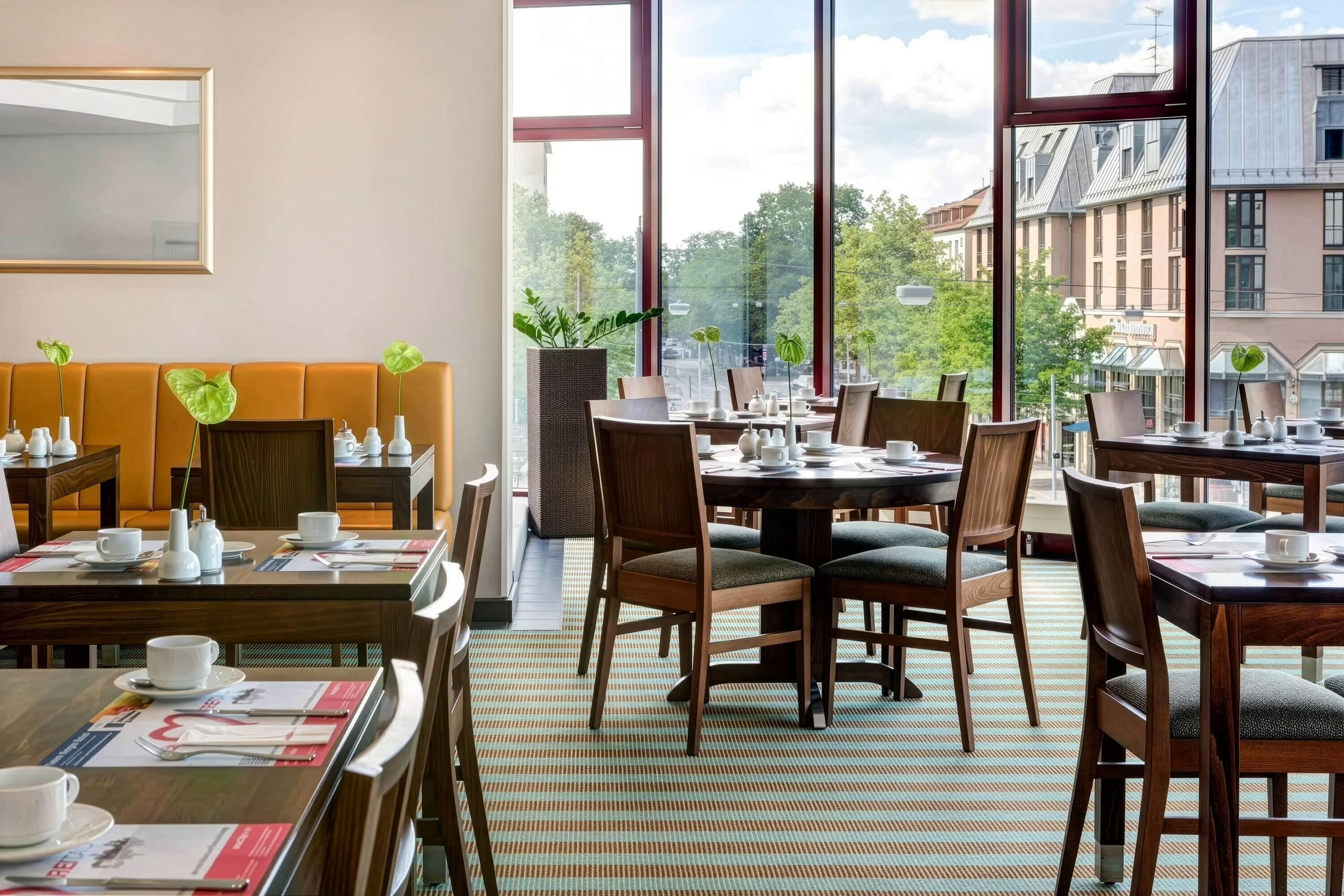 A hotel dining area with multiple tables and chairs, a sofa, and large windows offering a view of the cityscape.