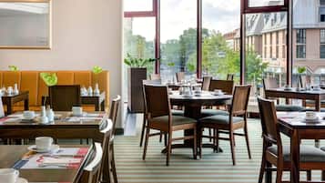 A hotel dining area with multiple tables and chairs, a sofa, and large windows offering a view of the cityscape.
