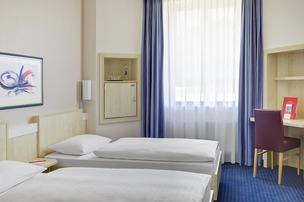 Habitación de hotel con dos camas, un escritorio, una silla, un armario y una ventana con cortinas.