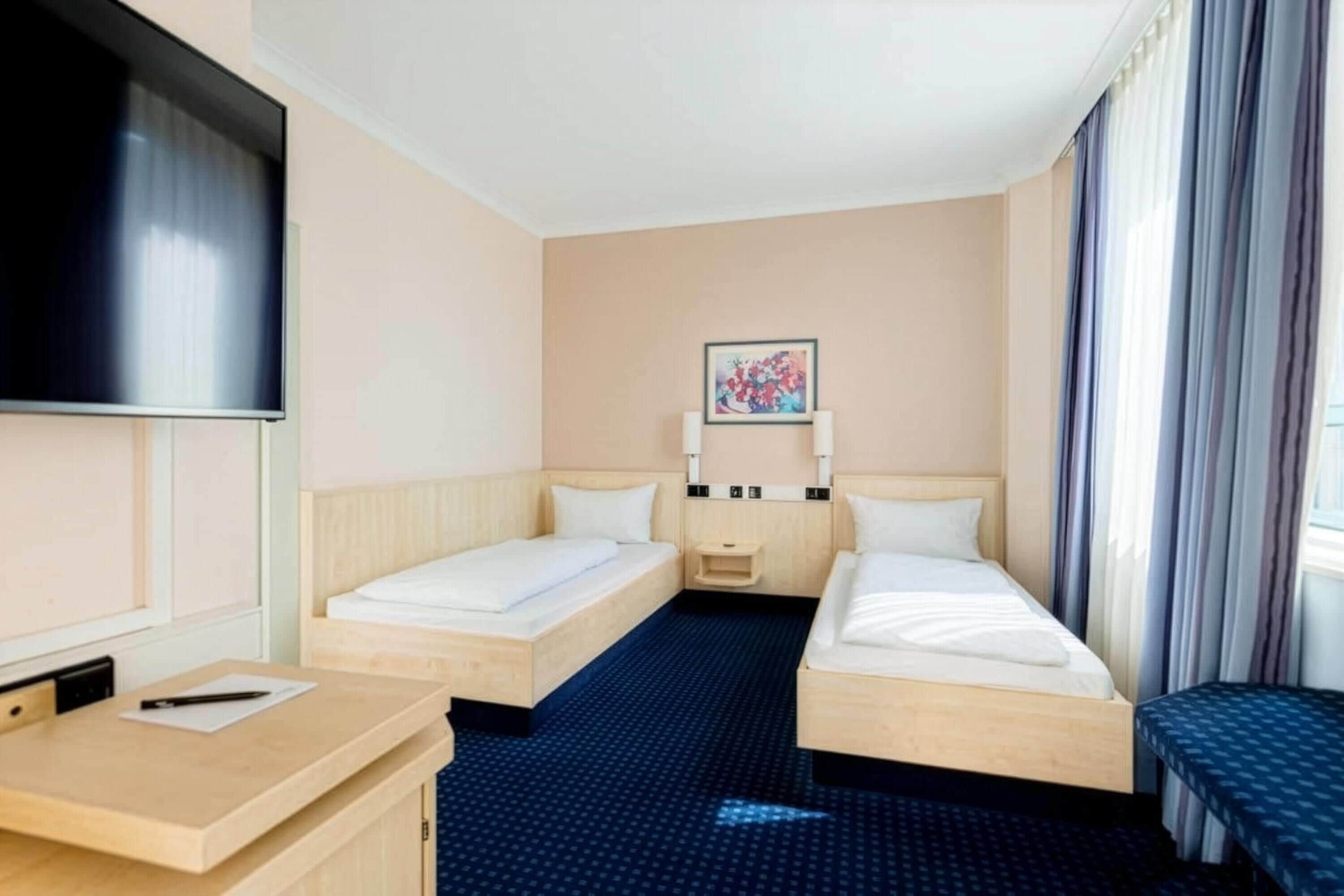 A hotel room with two single beds, a desk, a TV, a small bedside table, and a painting on the wall.