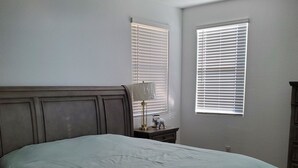 Un dormitorio con una cama, una mesita de noche, una lámpara y dos ventanas con persianas.