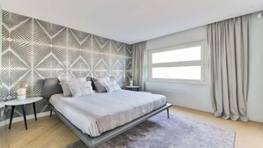 A modern bedroom with a geometric patterned wall, a large bed with a bench, a small round table, and a window with curtains. - QUAINT HIGH-FLOOR RETREAT ON QUAI D'ORSAY – EMBRACE THE PARISIAN LIFESTYLE (Paris)