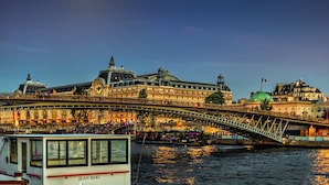 A historic building with a prominent clock tower, a green dome, and a bridge with people walking across it. - QUAINT HIGH-FLOOR RETREAT ON QUAI D'ORSAY – EMBRACE THE PARISIAN LIFESTYLE (Paris)