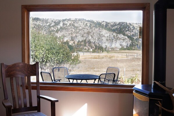 Ein Zimmer mit großem Fenster und Blick auf eine gebirgige Landschaft, ein Holzsessel, ein kleiner runder Tisch und ein schwarzer Heizkörper.