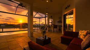 Amplia sala de estar con un gran ventanal que da a un cuerpo de agua y un atardecer. Se aprecia un sofá rojo, una mesa de centro con un jarrón, y un comedor con sillas y una mesa.