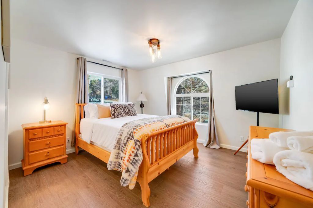 Un dormitorio con cama de madera, una mesita de noche, televisor y ventana con cortinas.