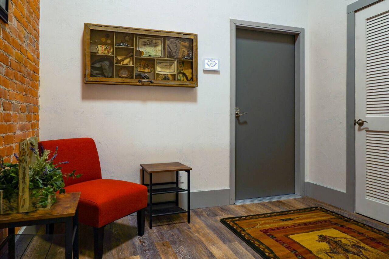 Une pièce avec un fauteuil rouge, une petite table en bois sur laquelle est posée une plante, une vitrine murale, une porte grise et un tapis à motifs.