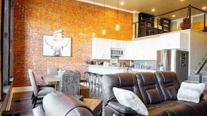 A modern living room with a brick wall, leather sofa, dining area, and a kitchen in the background. - West Empire Theatre Loft, Gorgeous Space on Main Street for 4! Local Golfing 50% Off! (Baker City)