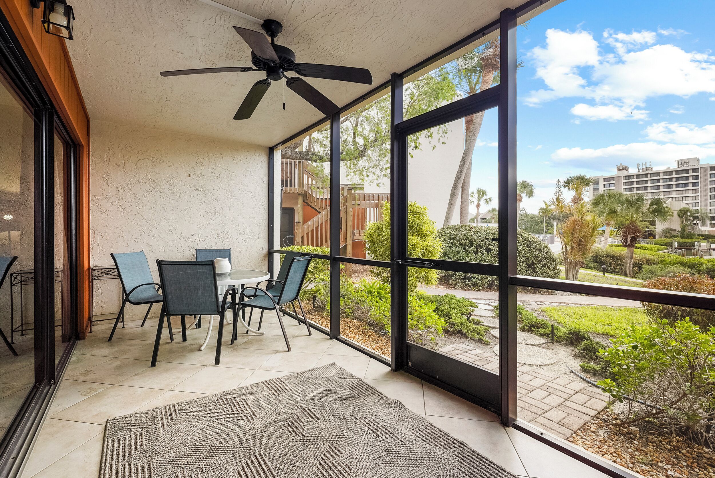A covered patio with a ceiling fan, a table with chairs, and a view of a landscaped garden and buildings in the background.