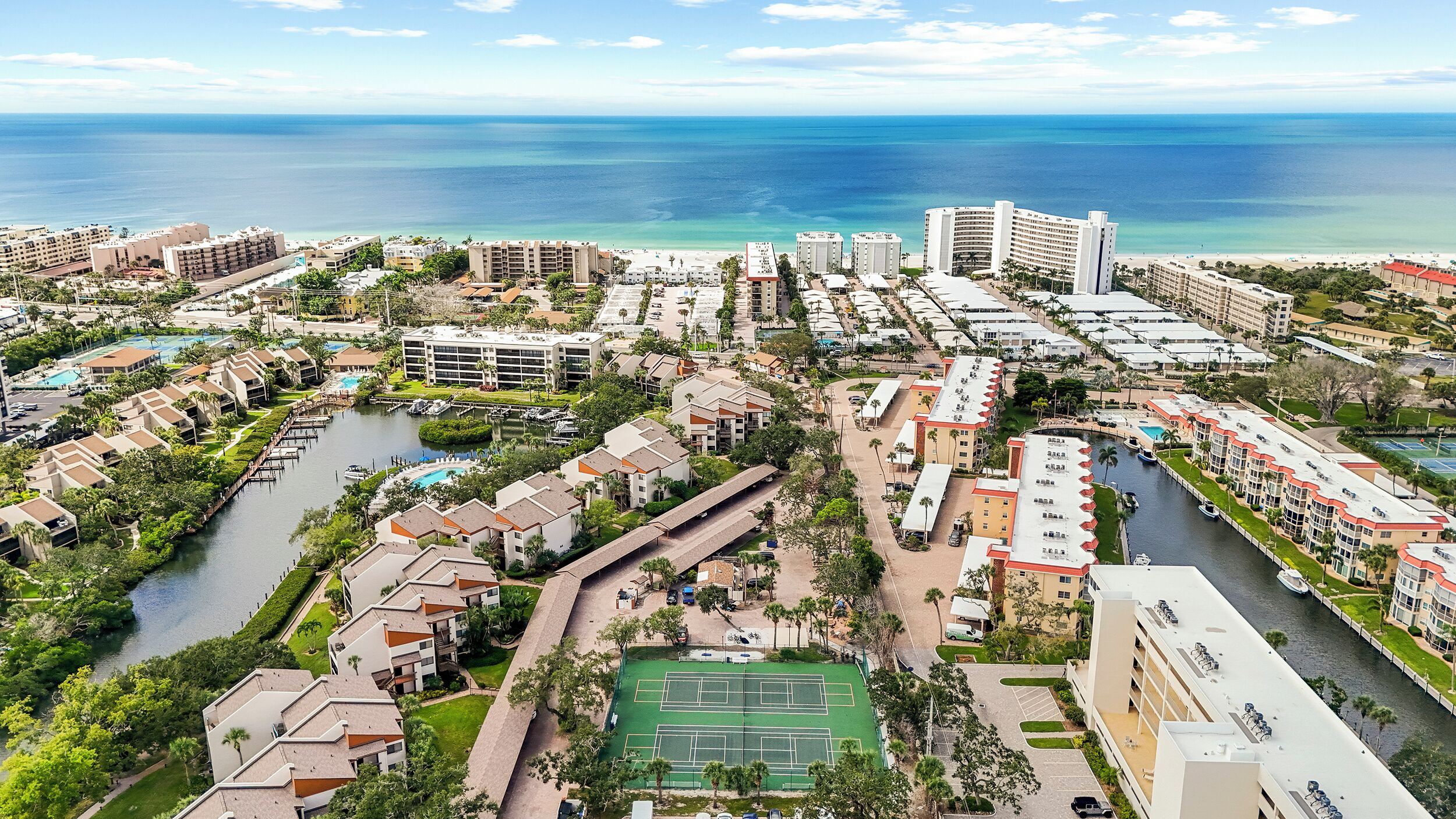 An aerial view of a coastal resort area with multiple buildings, a marina, and a tennis court.