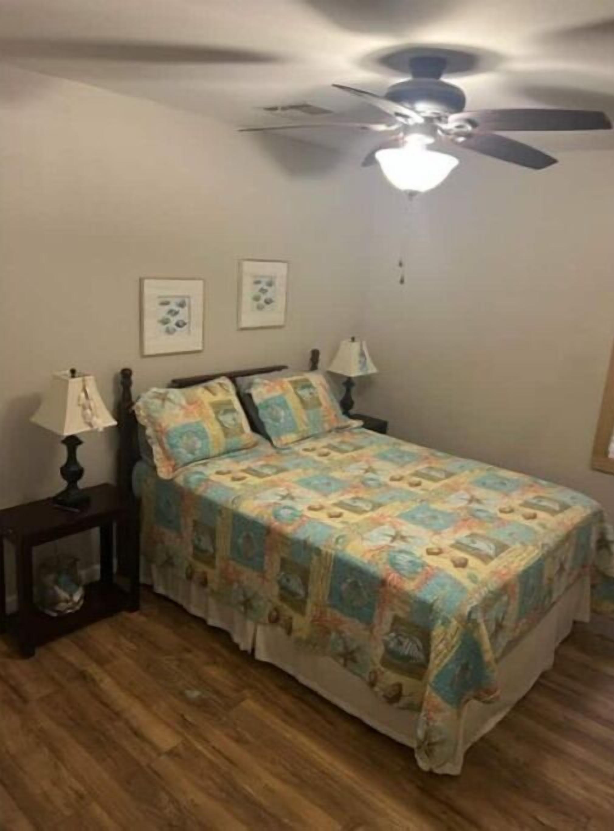 A bedroom with a bed, two bedside tables with lamps, a ceiling fan, and two framed pictures on the wall.