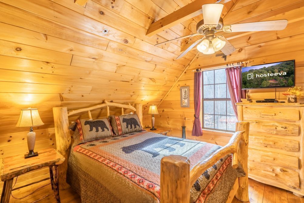 Un dormitorio rústico de cabaña de madera con una cama, cabecera de madera, ventilador de techo, televisor y ventana con cortinas.