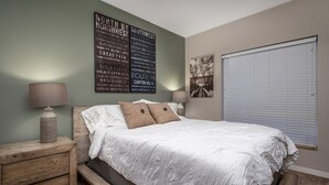 A bedroom with a bed, two bedside tables, a wall-mounted lamp, and a large window with blinds.