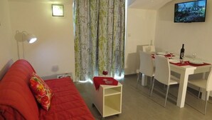 A compact living space with a red sofa, a small white table with a bottle and glasses, and a TV mounted on the wall.