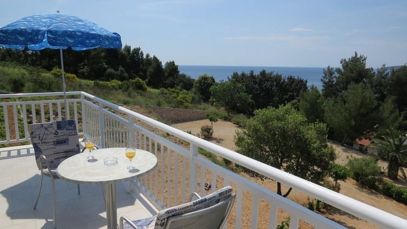 Een balkon met een tafel, stoelen, een parasol en uitzicht op zee en groen.