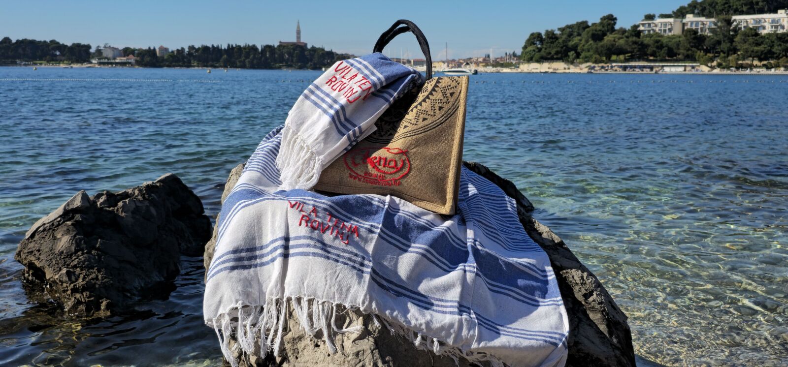 Uma toalha e uma bolsa sobre uma rocha perto de um lago límpido, com edifícios ao fundo.