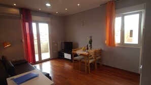 Ampla sala de estar com piso de madeira, mesa de jantar com cadeiras, TV de tela plana e porta de vidro deslizante que dá para uma varanda.