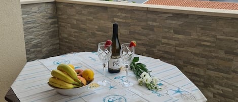 A table set on a balcony with a stone wall backdrop, a bottle of wine, glasses, and a bowl of fruit.