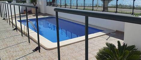 A rooftop swimming pool with a view of the sea, surrounded by palm trees and a tiled deck.