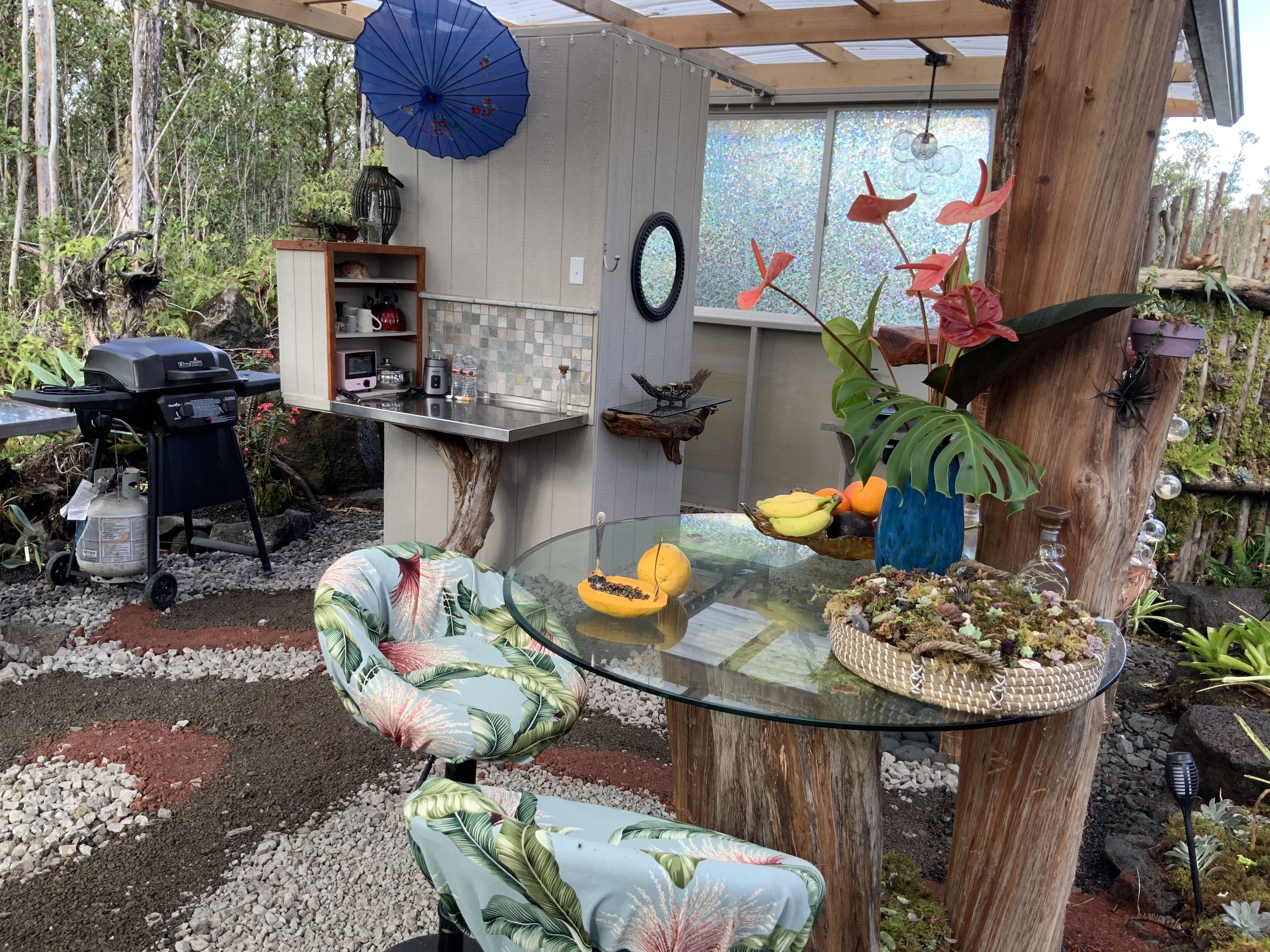 Une table en verre, une chaise au style tropical, un parasol bleu et une structure en bois rustique.