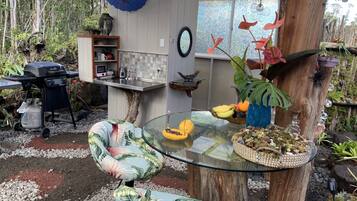 Une table en verre, une chaise au style tropical, un parasol bleu et une structure en bois rustique.