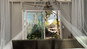 A bedroom with a bed, a wicker headboard, sheer curtains, a glass door leading to an outdoor area with plants and a rock formation. - Exotic garden cottage  at amazing volcanic fissure (Mountain View)