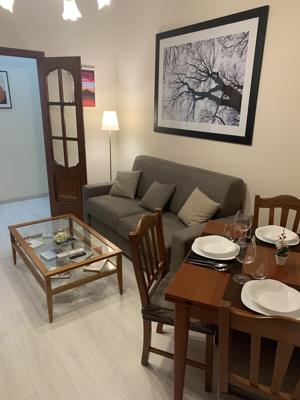 Een woonkamer met een grijze bank, een glazen salontafel, een eettafel gedekt met borden en wijnglazen, en een lijst met daarin een afbeelding van een boom.