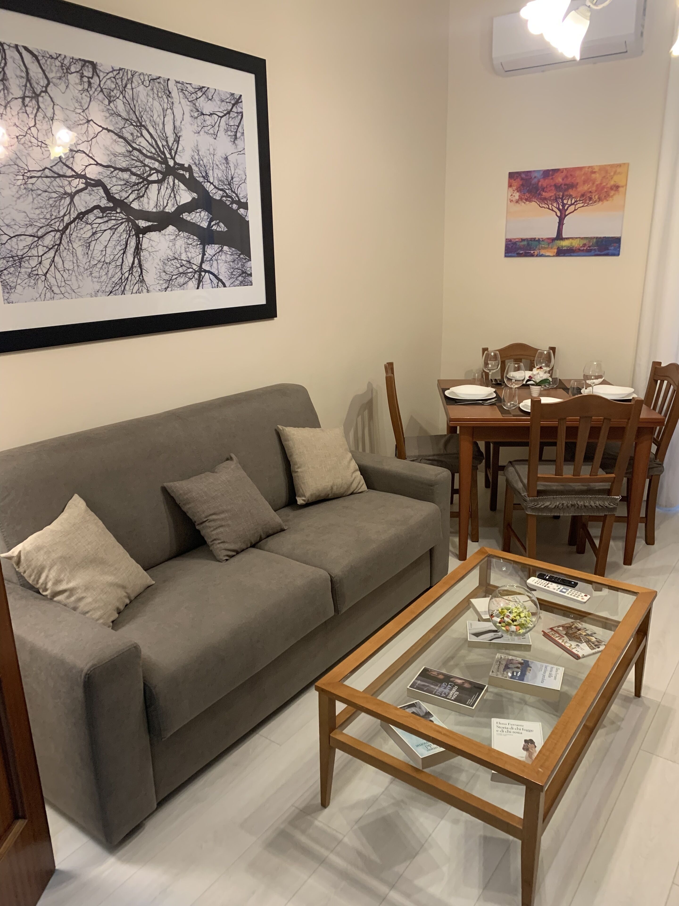 Un salon avec un canapé gris, une table basse en verre sur laquelle sont posés des magazines, une table à manger avec des chaises, et deux œuvres d’art encadrées accrochées au mur.