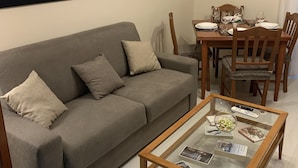 Un salon avec un canapé gris, une table basse en verre sur laquelle sont posés des magazines, une table à manger avec des chaises, et deux œuvres d’art encadrées accrochées au mur.