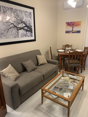 Uma sala de estar com um sofá cinza, uma mesa de centro de vidro com revistas, uma mesa de jantar com cadeiras e duas obras de arte emolduradas na parede.