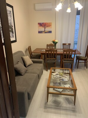 Een woonkamer met een bank, een glazen salontafel met tijdschriften, een eettafel met stoelen en een schilderij aan de muur.