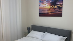 Un lit bien fait, avec du linge de lit blanc, une tête de lit grise et un tableau accroché au mur représentant un coucher de soleil sur l’océan.